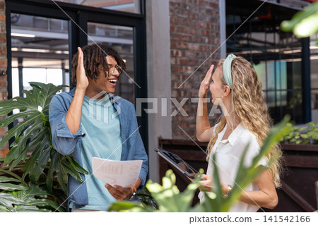 Diverse coworkers high-fiving while exchanging chart sheets and tablet in modern office with plants Diverse coworkers high-fiving while exchanging chart sheets and tablet in modern office with plants 141542166