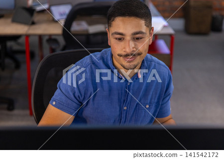 Man sitting on mesh chair at desk in modern open-plan office facing computer monitor, typing Man sitting on mesh chair at desk in modern open-plan office facing computer monitor, typing 141542172
