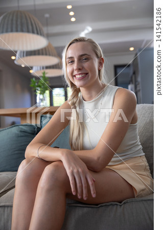 Woman sitting on in living area facing camera with wooden dining table in background Woman sitting on in living area facing camera with wooden dining table in background 141542186