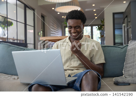 African american man celebrating success on laptop while sitting on sofa at home with smartphone African american man celebrating success on laptop while sitting on sofa at home with smartphone 141542190