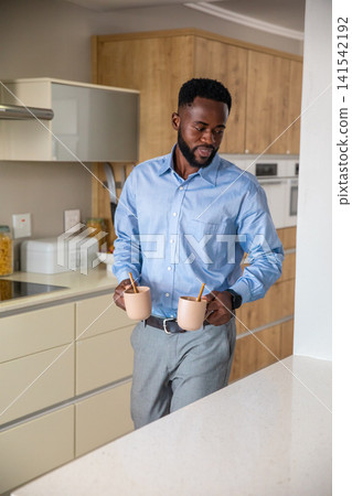 African american man holding ceramic mugs with wooden stirrers in home kitchen in blue shirt 141542192