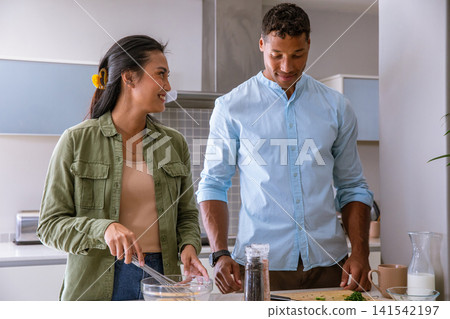 Diverse couple stirring batter in glass bowl and chopping herbs on cutting board at kitchen counter Diverse couple stirring batter in glass bowl and chopping herbs on cutting board at kitchen counter 141542197