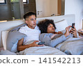 Diverse male partners lying under duvet using smartphones near wooden bedside table in bedroom 141542213