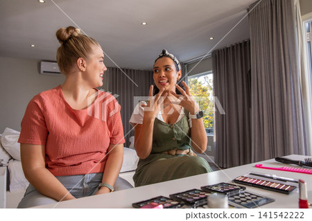 Diverse female friends discussing makeup tips at bedroom desk with palettes, brushes, smartphone Diverse female friends discussing makeup tips at bedroom desk with palettes, brushes, smartphone 141542222
