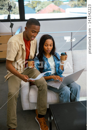 African American coworkers sitting on gray sofa in home office collaborating on laptop with coffee African American coworkers sitting on gray sofa in home office collaborating on laptop with coffee 141542223