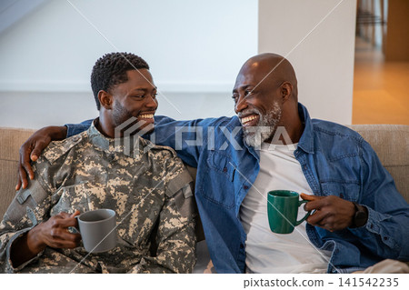 African american father and son smiling on couch at home with mugs, son wearing camouflage African american father and son smiling on couch at home with mugs, son wearing camouflage 141542235