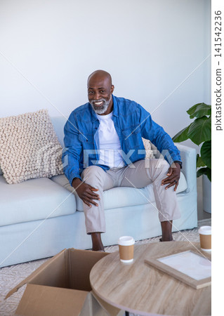 African american man sitting on sofa in living room leaning forward with coffee cups African american man sitting on sofa in living room leaning forward with coffee cups 141542236