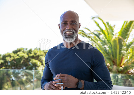 African american man standing on terrace holding coffee mug with navy shirt and smartwatch, ring 141542240