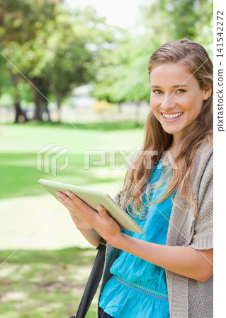 Adult female holding tablet and smiling in park, wearing blue blouse with bag strap, copy space Adult female holding tablet and smiling in park, wearing blue blouse with bag strap, copy space 141542272