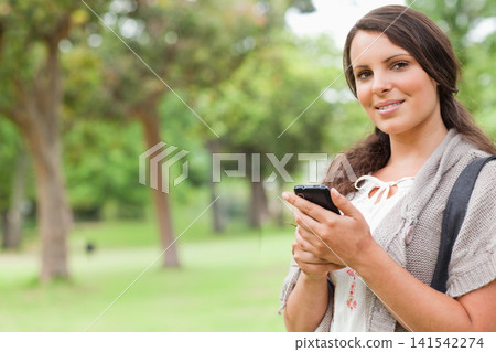 Teen woman standing in park using phone in tie-blouse cardigan and bag strap visible, copy space Teen woman standing in park using phone in tie-blouse cardigan and bag strap visible, copy space 141542274