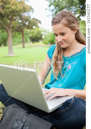 Female wearing teal top and jeans, sitting cross-legged on park grass using silver laptop, backpack Female wearing teal top and jeans, sitting cross-legged on park grass using silver laptop, backpack 141542277