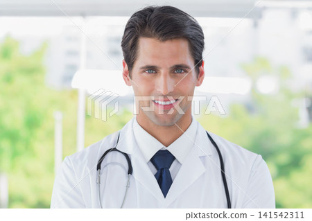Male doctor smiling and standing in clinic by large window with stethoscope and white coat Male doctor smiling and standing in clinic by large window with stethoscope and white coat 141542311