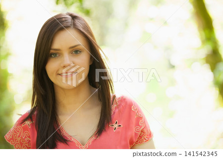 Adult woman standing in sunlit wooded area, wearing embroidered coral-pink blouse and smiling Adult woman standing in sunlit wooded area, wearing embroidered coral-pink blouse and smiling 141542345