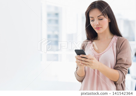 Female in cardigan standing in bright room near windows, holding phone in both hands, copy space Female in cardigan standing in bright room near windows, holding phone in both hands, copy space 141542364