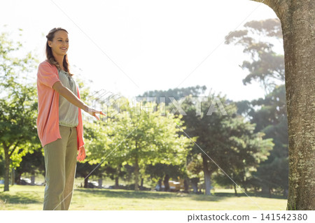 Woman standing on lawn in park wearing pink cardigan tan trousers near tree trunk, copy space Woman standing on lawn in park wearing pink cardigan tan trousers near tree trunk, copy space 141542380