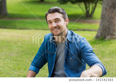Middle-aged man sitting on grassy lawn in park, wearing denim shirt over gray t-shirt and smiling Middle-aged man sitting on grassy lawn in park, wearing denim shirt over gray t-shirt and smiling 141542383