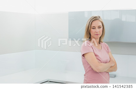 Middle-aged woman standing with arms crossed in kitchen near counter, sink and papers, copy space Middle-aged woman standing with arms crossed in kitchen near counter, sink and papers, copy space 141542385