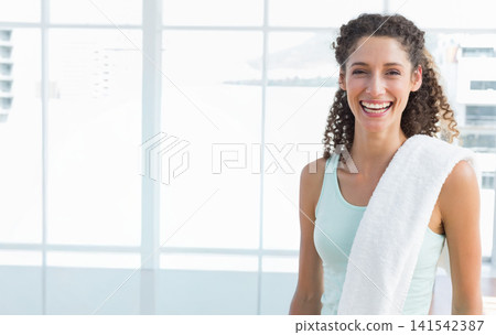 Female in twenties standing smiling by grid windows in bright fitness studio with towel, copy space Female in twenties standing smiling by grid windows in bright fitness studio with towel, copy space 141542387