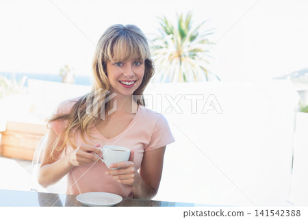 Female sitting at glass table on terrace holding white teacup, saucer, wearing pink top, smiling Female sitting at glass table on terrace holding white teacup, saucer, wearing pink top, smiling 141542388
