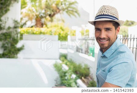 Man leaning on black railing on sunlit terrace, wearing straw hat and blue shirt, copy space 141542393