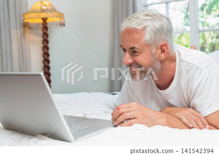 Senior man lying on bed propped on elbows, looking at silver laptop and wearing wedding band Senior man lying on bed propped on elbows, looking at silver laptop and wearing wedding band 141542394