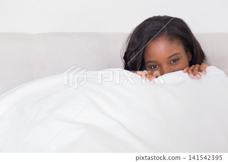 African American woman peeking from white duvet on bed, gripping duvet near headboard, copy space African American woman peeking from white duvet on bed, gripping duvet near headboard, copy space 141542395