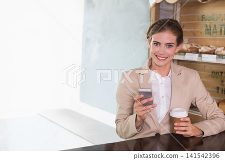 Adult female in beige blazer sitting at cafe counter holding phone, to-go cup near bakery shelves Adult female in beige blazer sitting at cafe counter holding phone, to-go cup near bakery shelves 141542396