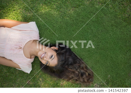 Asian adult woman lying on green lawn smiling, spreading long wavy hair, wearing pink blouse 141542397
