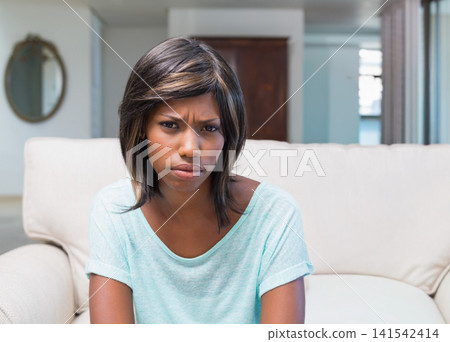 African American woman sitting on beige sofa in living room, frowning and holding cushion African American woman sitting on beige sofa in living room, frowning and holding cushion 141542414