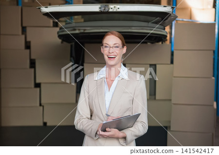 Female manager wearing beige blazer holding clipboard under open trunk lid in warehouse with boxes Female manager wearing beige blazer holding clipboard under open trunk lid in warehouse with boxes 141542417