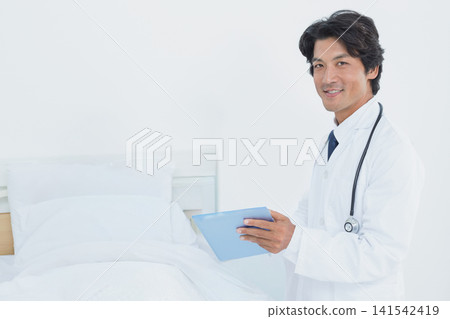 Japanese male doctor in lab coat and stethoscope reading tablet beside medical bed, copy space Japanese male doctor in lab coat and stethoscope reading tablet beside medical bed, copy space 141542419