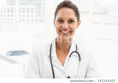 Mid-adult female doctor smiling at camera, wearing white coat and stethoscope in clinic by windows Mid-adult female doctor smiling at camera, wearing white coat and stethoscope in clinic by windows 141542423
