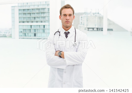 Male physician standing centered in bright clinic with tall windows, wearing lab coat, stethoscope 141542425