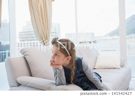 Child girl wearing navy dress reclining on sofa at home, resting chin and gazing through window Child girl wearing navy dress reclining on sofa at home, resting chin and gazing through window 141542427