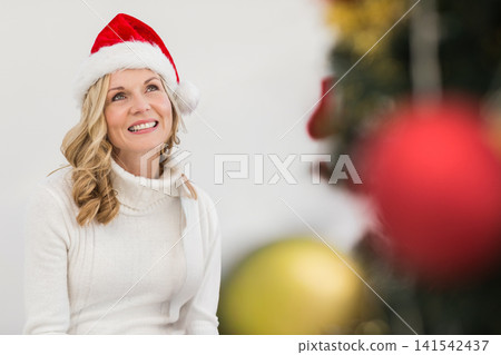 Middle-aged woman posing in Santa hat and turtleneck in studio, smiling at red baubles, copy space Middle-aged woman posing in Santa hat and turtleneck in studio, smiling at red baubles, copy space 141542437