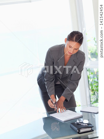 Female exec grey suit leaning over glass desk at office, marking papers with pen, notebook nearby Female exec grey suit leaning over glass desk at office, marking papers with pen, notebook nearby 141542454