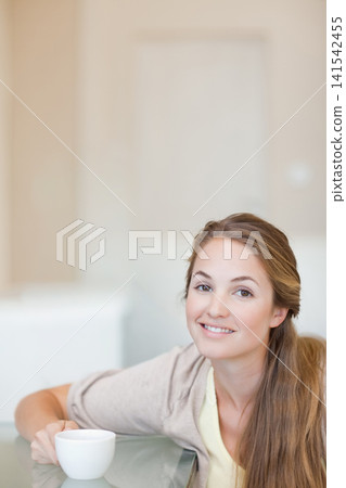 Adult female leaning on glass table at home wearing light sweater, holding ceramic cup, copy space Adult female leaning on glass table at home wearing light sweater, holding ceramic cup, copy space 141542455