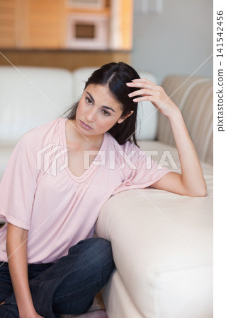 Woman sitting on floor beside cream leather sofa touching hair in living room, wearing pink top Woman sitting on floor beside cream leather sofa touching hair in living room, wearing pink top 141542456