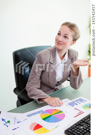 Female professional wearing blazer sitting at desk gesturing while reviewing charts and keyboard Female professional wearing blazer sitting at desk gesturing while reviewing charts and keyboard 141542457