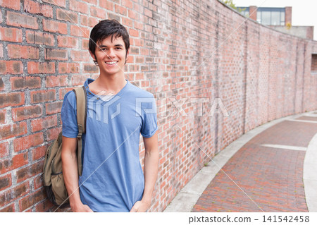 Asian teen leaning against red-brick wall on brick path in V-neck with khaki backpack, copy space Asian teen leaning against red-brick wall on brick path in V-neck with khaki backpack, copy space 141542458