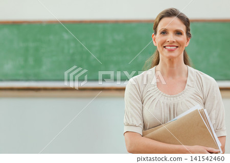 Woman teacher holding papers, tan folder by green board classroom front with chalk tray, copy space Woman teacher holding papers, tan folder by green board classroom front with chalk tray, copy space 141542460