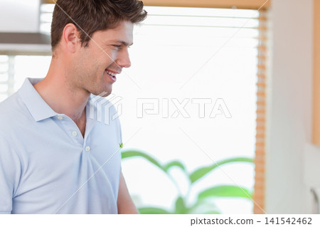 Man in 30s smiling looking right at home wearing light-blue polo, plant, blinds, copy space Man in 30s smiling looking right at home wearing light-blue polo, plant, blinds, copy space 141542462