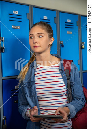 Teenage girl wearing denim jacket using tablet in school hall by lockers with backpack Teenage girl wearing denim jacket using tablet in school hall by lockers with backpack 141542473