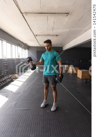 Man lifting hex dumbbells in sportswear on mat in gym weight area under fluorescent lights Man lifting hex dumbbells in sportswear on mat in gym weight area under fluorescent lights 141542479