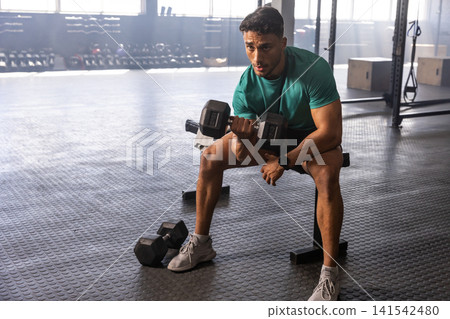 Man gripping heavy dumbbell, performing seated bicep curl on flat bench in gym free-weight area Man gripping heavy dumbbell, performing seated bicep curl on flat bench in gym free-weight area 141542480