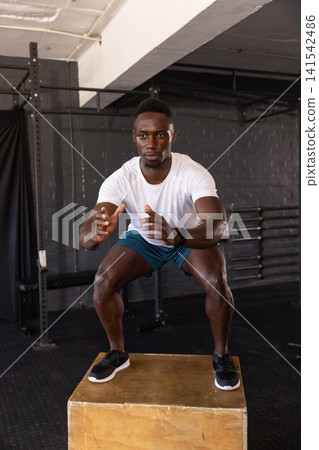 African American man doing plyometric box jump at gym with wooden box, squat rack and smartwatch African American man doing plyometric box jump at gym with wooden box, squat rack and smartwatch 141542486