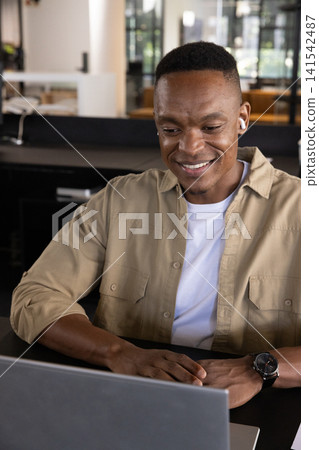African american man working on laptop at open-plan office wearing wireless earbuds, wristwatch African american man working on laptop at open-plan office wearing wireless earbuds, wristwatch 141542487