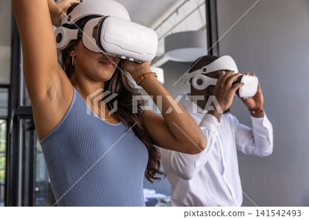 Diverse coworkers adjusting white vr headsets in office with ceiling ducts, glass partitions Diverse coworkers adjusting white vr headsets in office with ceiling ducts, glass partitions 141542493