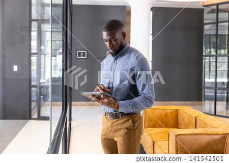 African American man wearing business casual standing in lounge near glass partition holding tablet African American man wearing business casual standing in lounge near glass partition holding tablet 141542501