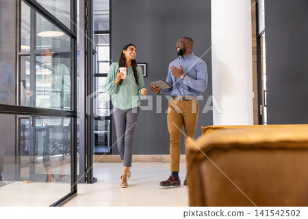 Diverse coworkers walking through office corridor in business attire holding coffee cup, tablet 141542502